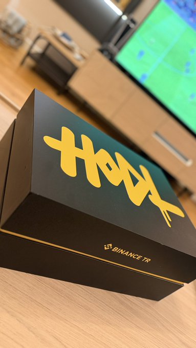 A sleek black gift box with bold yellow "XOX" lettering and a "BINANCE TR" label sits on a wooden surface, with a TV displaying a soccer match blurred in the background—matching the post's sentiment of being delighted by thoughtfully prepared gifts from Binance Turkey.