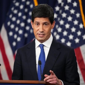 A man in a dark suit with a blue polka-dot tie speaks at a podium in front of multiple American flags.