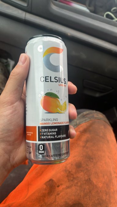 A hand holds a can of CELSIUS Sparkling Mango Lemonade Flavored energy drink, which is labeled as zero sugar and zero calories, inside a vehicle.