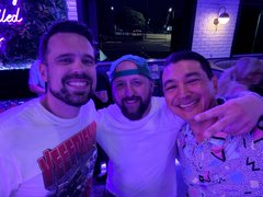 Three men smile in a selfie inside a karaoke bar bathed in purple lighting. The image ties to the post's call for the VeeFriends community to leave positive reviews for the karaoke venue they enjoyed.