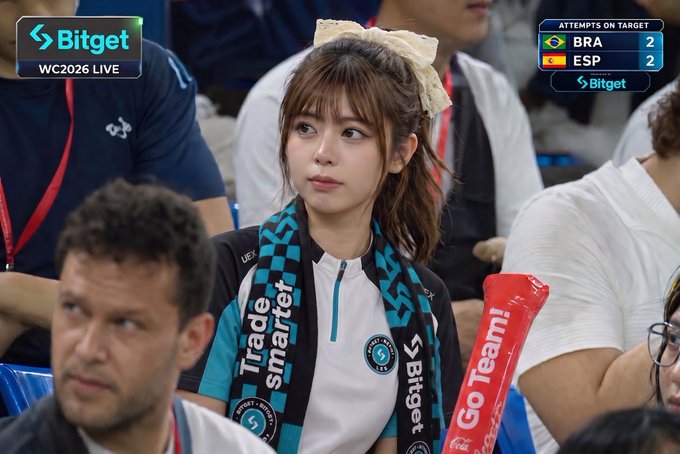 The image shows a woman wearing a black and white striped scarf, sitting among other people at a sports event. The crowd appears to be engaged in watching the game, with some of them holding red flags or banners. The focus of the image is on the woman's scarf, which stands out against the background. The post about BitgetTC seems unrelated to the scene and does not provide any information about soccer or the event taking place at the stadium.