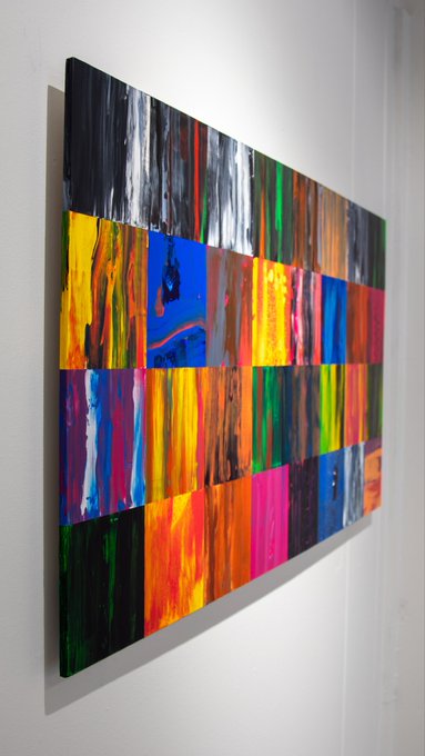 An abstract rectangular canvas painting composed of a grid of colorful squares in vibrant hues of red, yellow, blue, green, and black, mounted on a white wall.