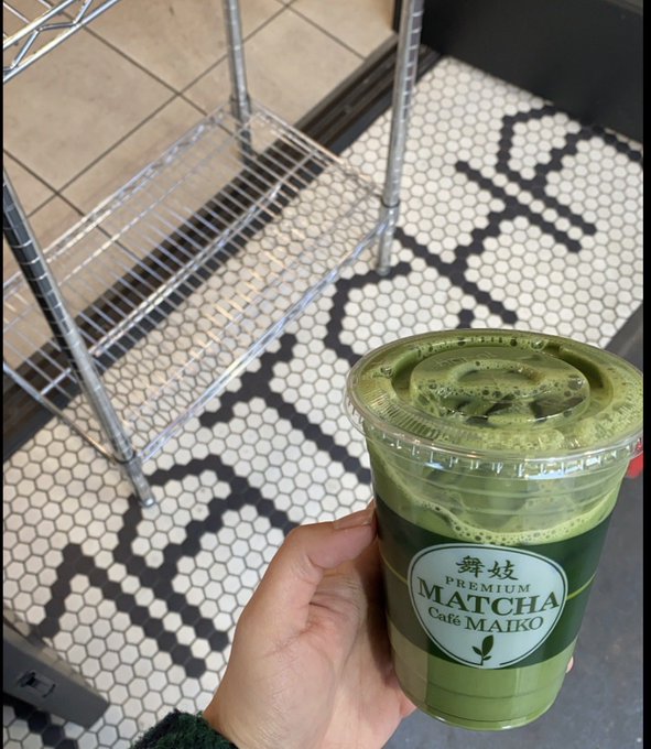 Hand holding a plastic cup of iced matcha from 'Premium Cafe MAIKO' in a cafe with a black and white hexagonal tiled floor.
