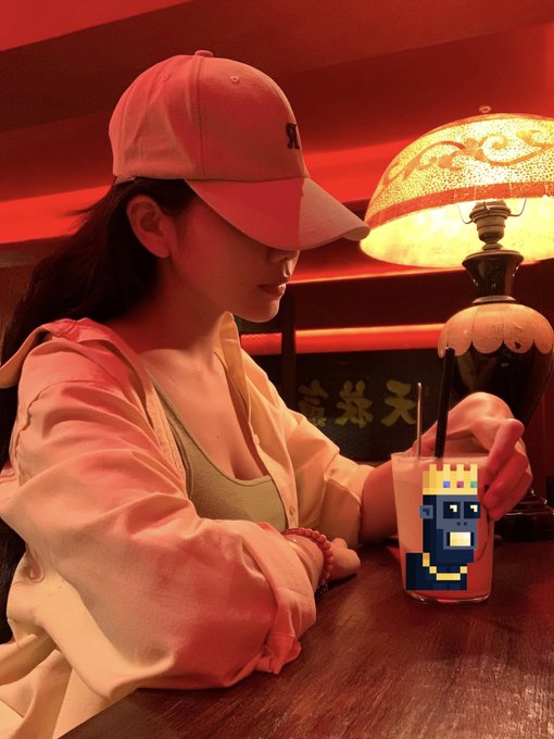 A young woman wearing a pink cap and light-colored jacket sits at a wooden table in a warmly lit red-hued room, with a pixel-art king character overlaid on the cup in front of her.