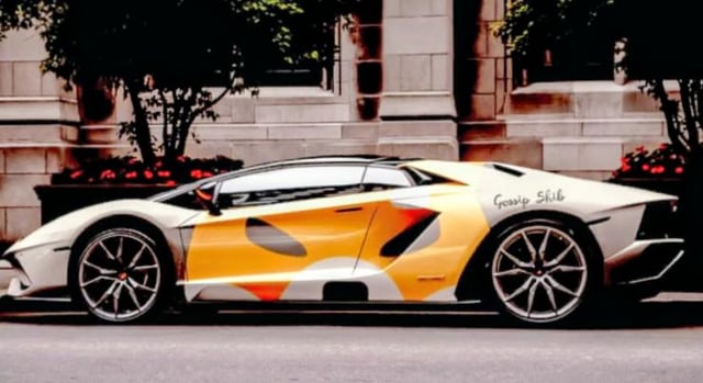 A side-profile shot of a white and orange camouflage-patterned Lamborghini supercar with "Gossip Slut" text on the rear quarter panel, parked near a building.