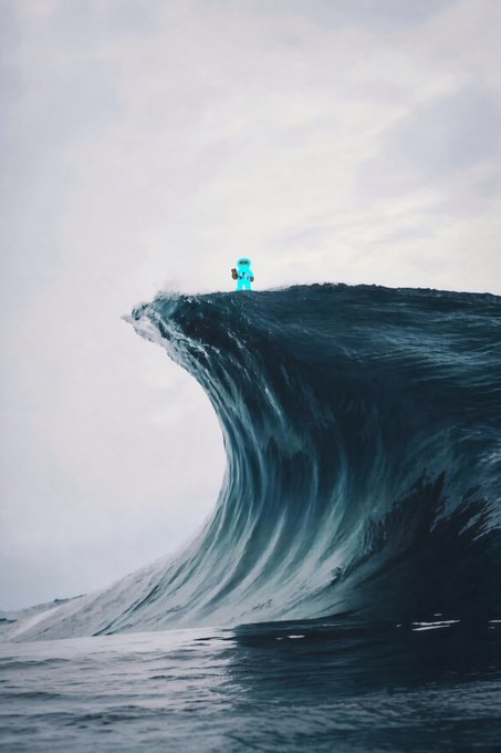 A small blue Minecraft-style character stands atop a towering, curling ocean wave against a pale, cloudy sky.
