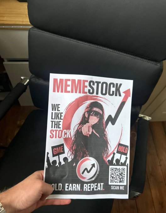 A hand holds up a "MEMESTOCK" promotional flyer featuring an anime-style figure, "WE LIKE THE STOCK" text, "GME" and "HOLD" protest signs, a rising arrow, and a "SCAN ME" QR code, echoing the post's bullish pitch for the $MEMESTOCK token.