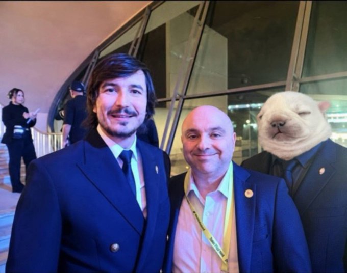 Two men in dark suits pose for a photo at what appears to be a formal event, with one notably wearing a large white cat mask, alongside a partial yellow lanyard visible on the middle figure. The "free von piblo" caption suggests a playful reference, possibly alluding to Pablo (perhaps a name reference), with the masked figure adding to the humorous, surreal tone of the image.