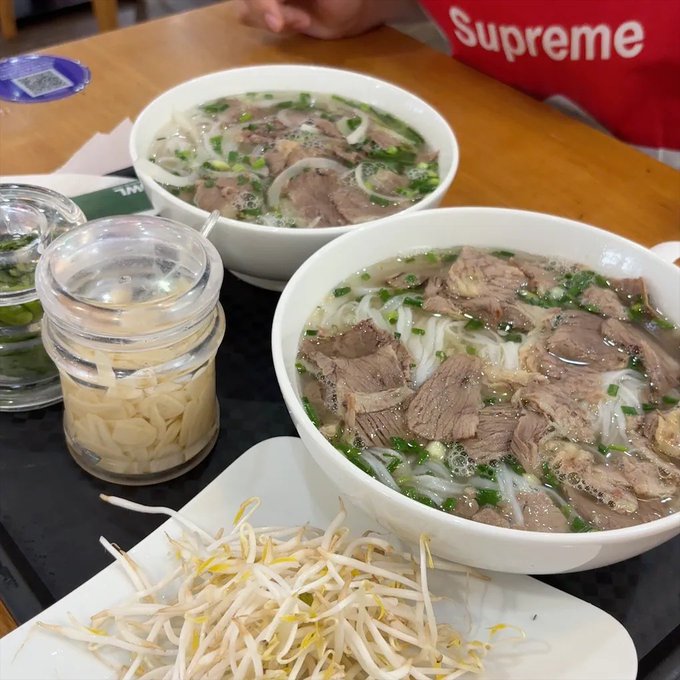 Two bowls of Vietnamese beef pho garnished with onions and scallions sit on a wooden table alongside plates of fresh bean sprouts and jars of sliced garlic, with a person wearing a red "Supreme" t-shirt in the background. The meal reflects a moment of pause and reflection, complementing the post's contemplation about being mentally scattered and uncertain about which direction (crypto, Web 3, or lifestyle) to focus on next.