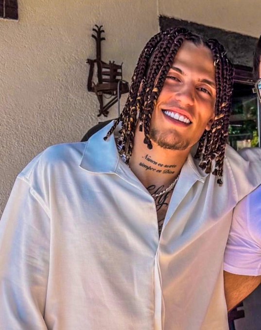 A smiling young man with long braided hair, a goatee, and a neck tattoo reads 'Nunca es suerte siempre es Dios' while wearing an open white satin shirt.