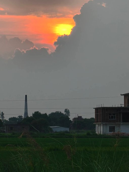 A photograph of a sunset with vibrant orange and yellow clouds over a rural landscape, featuring a smoking brick chimney, a partially constructed multi-story building, trees, and a field of green crops in the foreground.