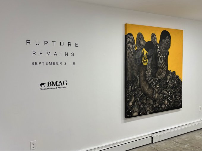 An art gallery wall displays the text 'RUPTURE REMAINS SEPTEMBER 2-8 BMAG Bitcoin Museum & Art Gallery' next to a large painting featuring black bird-like figures and yellow accents on a dark background.