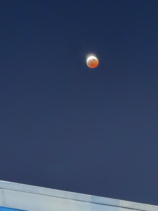 A reddish-orange moon, likely a blood moon during a lunar eclipse, captured against a dark blue night sky above a building's roofline.