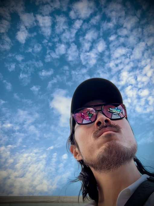 A low-angle portrait of a man wearing a black cap and sunglasses reflecting a landscape, set against a blue sky with scattered clouds.