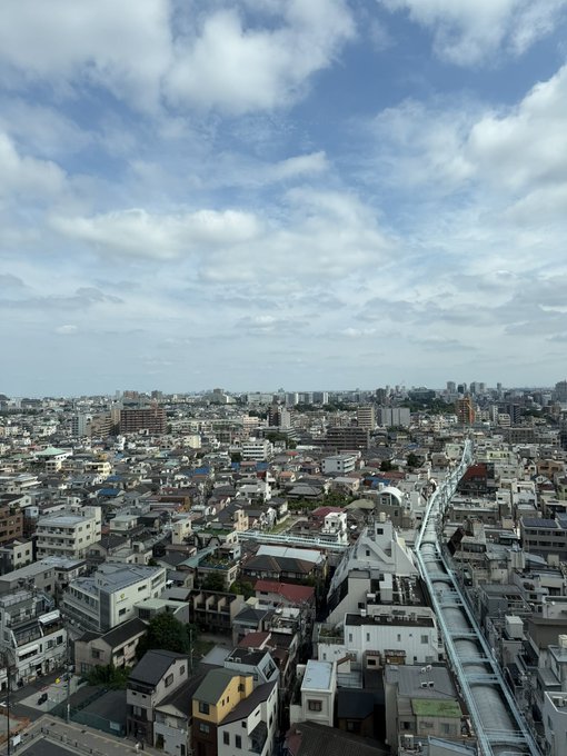 An elevated view of a sprawling cityscape, likely a Japanese urban area, featuring dense residential and commercial buildings under a partly cloudy blue sky.