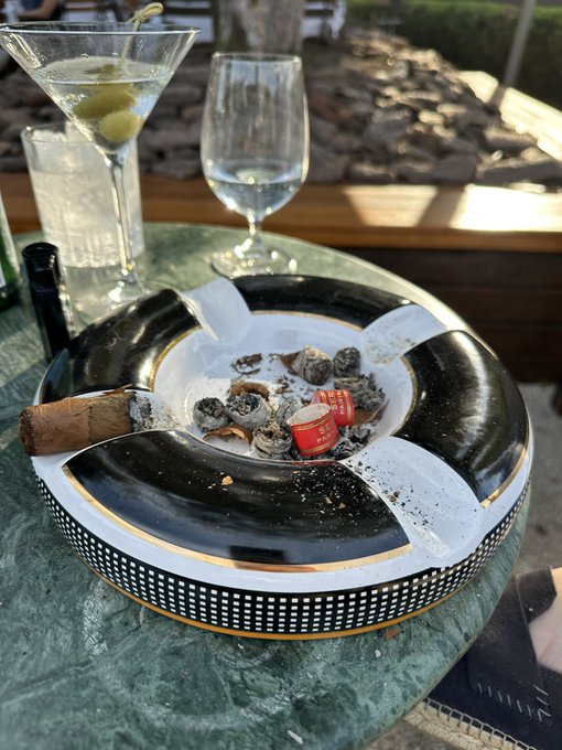 A large black and white ashtray overflowing with ash, cigarette butts, and empty packs sits on a green marble outdoor table next to a martini with olives and an empty wine glass.