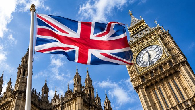 The British Union Jack flag waving in the wind with Big Ben's clock tower visible in the background against a blue sky.