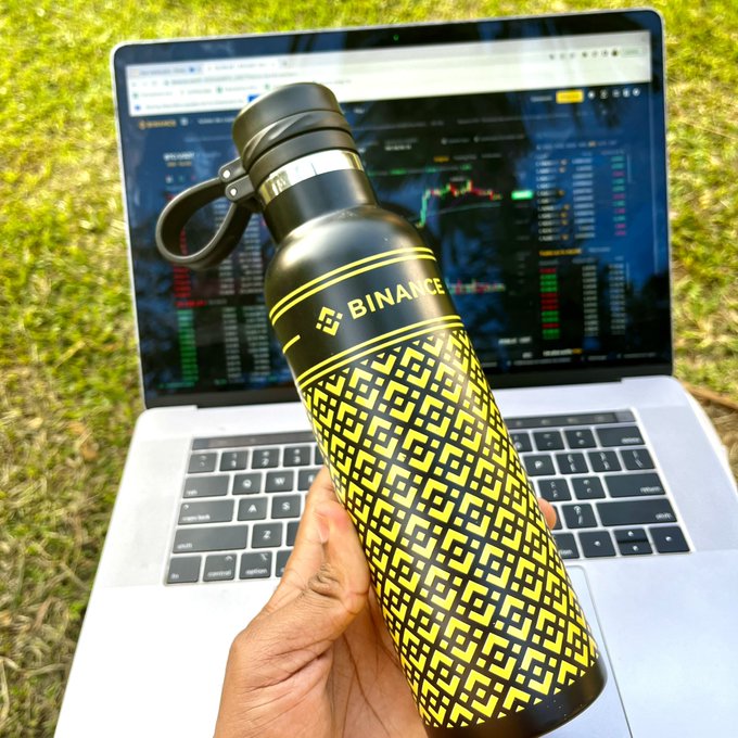 The image features a person holding up a black and yellow water bottle, which has the word "BINANCE" printed on it. The person's hand is positioned over a laptop computer that is open and displaying a screen filled with financial data. The background of the image shows a grassy area where the person is standing outdoors.