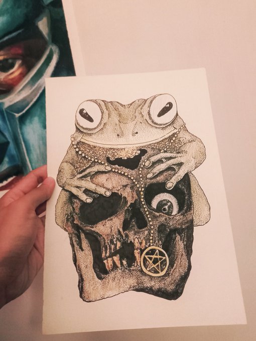 A hand holds up a black and white pointillism-style illustration of a frog perched on a human skull, adorned with jewelry including a beaded necklace and a pentagram pendant.