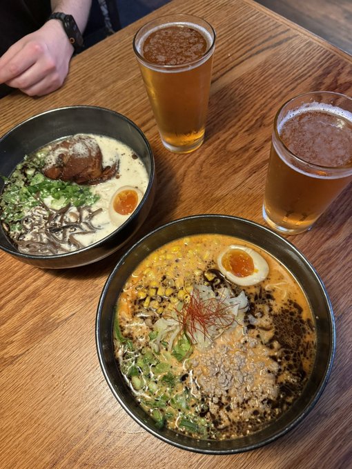 Two bowls of ramen with various toppings like mushrooms, corn, soft-boiled egg, and green onions sit on a wooden table next to two glasses of beer.