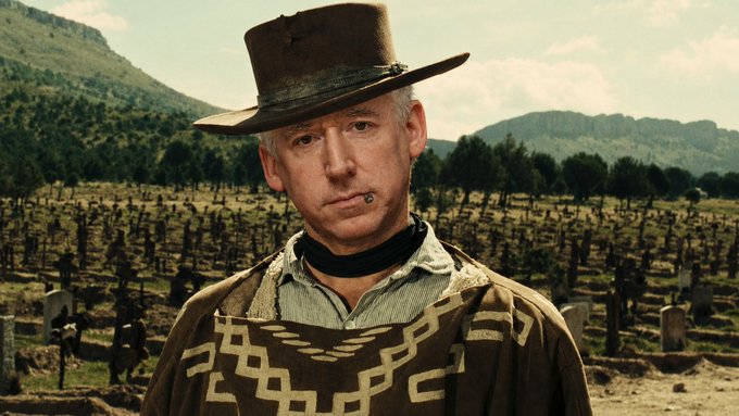 A man wearing a brown cowboy hat and a southwestern-patterned poncho, smoking a cigarette, standing in front of a cemetery and mountainous landscape.