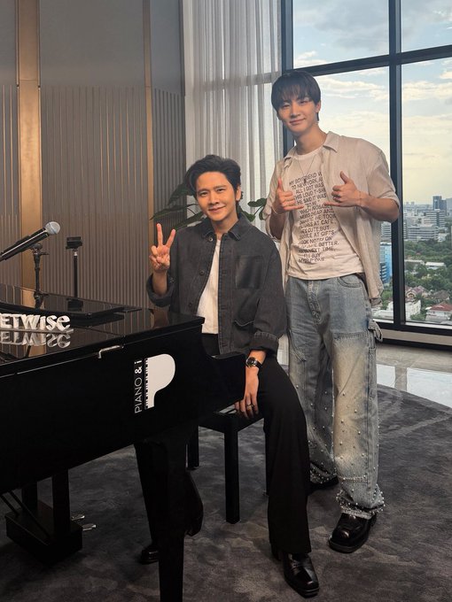 Two men pose in a high-rise room with large windows; one sits on a bench near a piano marked "PIANO & I" and "NETWISE THEATRE," while the other stands wearing a white t-shirt with printed text.