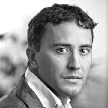 A black-and-white headshot of a man with short curly hair wearing a light-colored suit jacket and white shirt, looking slightly off-camera.