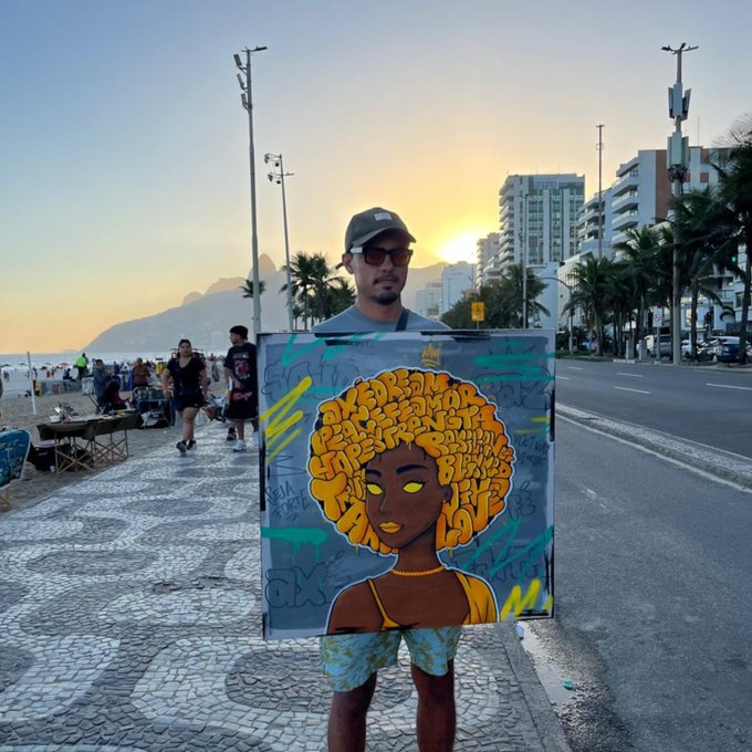 A man wearing a cap stands on a beachfront promenade at sunset, holding a colorful graffiti-style painting of a woman with an afro and yellow eyes, with palm trees and city buildings in the background.