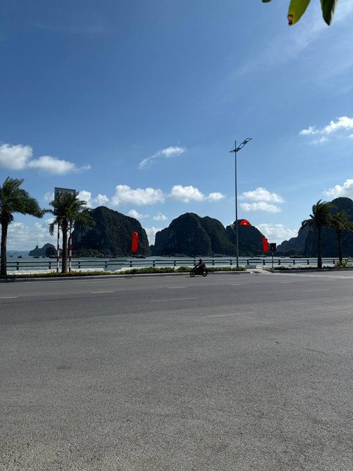 Scenic coastal road view featuring limestone karst mountains, palm trees, and a person riding a motorbike under a blue sky with scattered clouds.