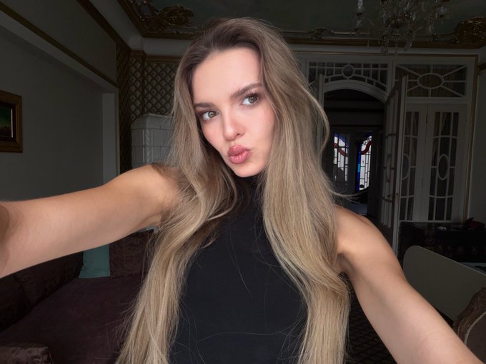A blonde woman in a black sleeveless top takes a selfie, pursing her lips in a room with ornate details.