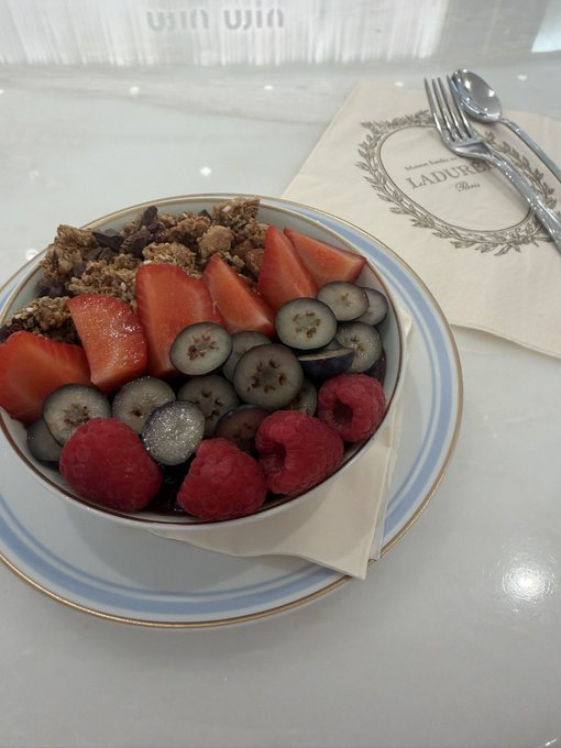 A bowl of granola topped with sliced strawberries, blueberries, and raspberries sits on a Ladurée-branded napkin beside silverware, presented as a clean, refreshing breakfast to accompany a motivational post about grinding through bear markets in crypto to capitalize on bull runs.