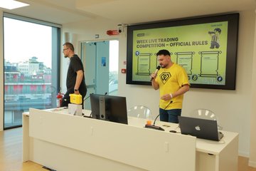 Two men stand behind a desk, with the man on the left holding a yellow toolbox and the other wearing a yellow t-shirt and microphone.