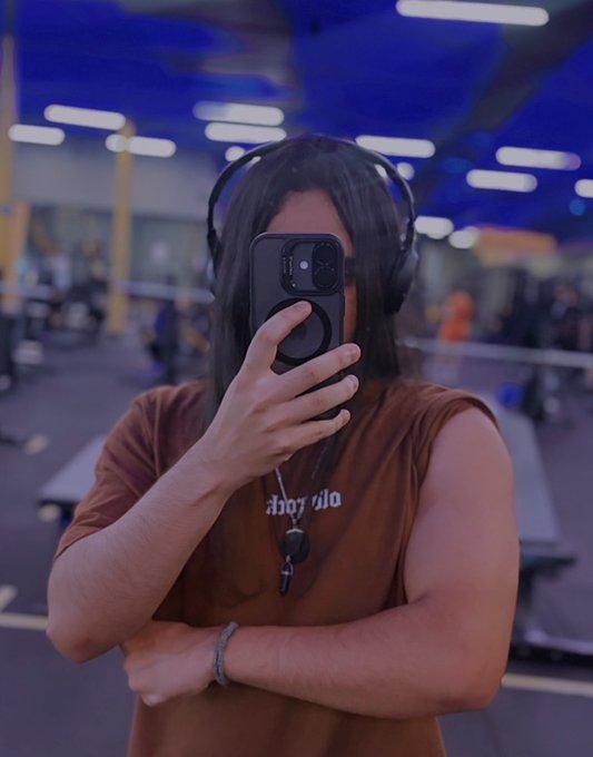 A woman wearing headphones takes a mirror selfie in a gym, partially covering her face with a black smartphone.