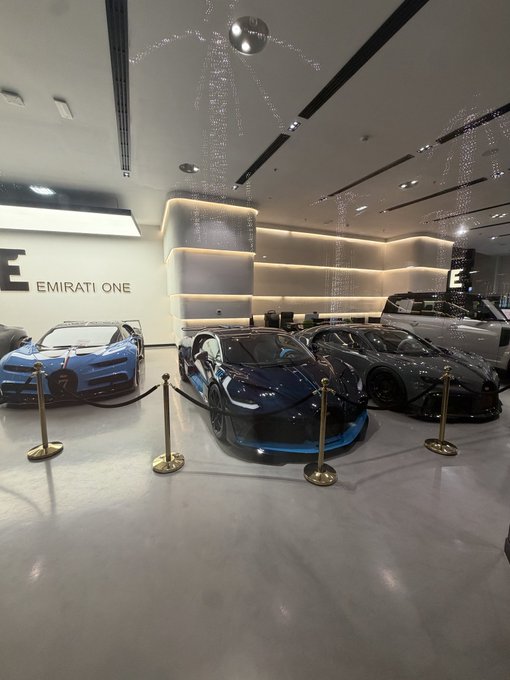 A lineup of sleek Bugattis—including a blue Chiron and a black model—on display at the "EMIRATI ONE" showroom, where stanchions cordon off the exotic hypercars under cascading lights. The image evokes the post's promise of life-changing wealth, visualizing the ultra-luxury lifestyle that awaits when BTC, SOL, and ETH reach their projected highs.