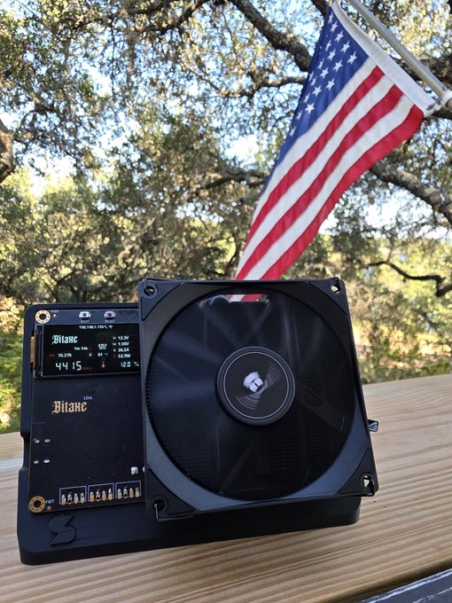The image showcases the Bitaxe Naja Duo, the first BM1373 Bitcoin home miner assembled in the USA, featuring a digital display reading "44.15" and "Bitaxe" branding, displayed on a wooden surface with an American flag waving in the background to highlight its domestic manufacturing.