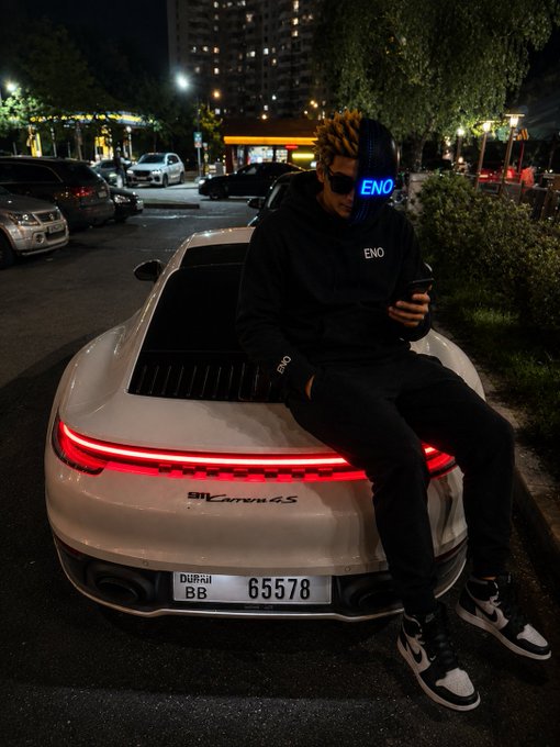 A person in a black "ENO" hoodie and pixelated mask sits on the back of a white 911 Carrera 4S with Dubai plate 65578, illuminated by its red taillights at night. The caption "Good night CT" matches the nighttime farewell vibe of winding down after a long day's progress.