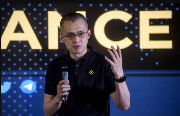 Binance CEO Changpeng Zhao (CZ) speaks into a microphone while wearing a black Binance polo shirt and glasses, gesturing with his hand in front of a large illuminated 'BINANCE' logo backdrop.