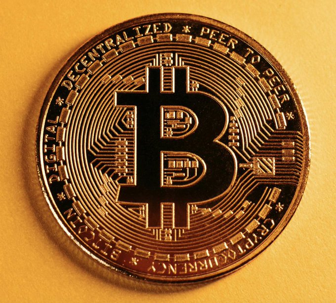 A close-up photo of a physical Bitcoin coin with the prominent 'B' logo and the inscription 'DECENTRALIZED PEER TO PEER' and 'DIGITAL CURRENCY'.