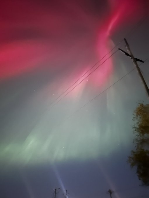 Pink and green aurora lights glow across a starry night sky, crossed by power lines and a utility pole.