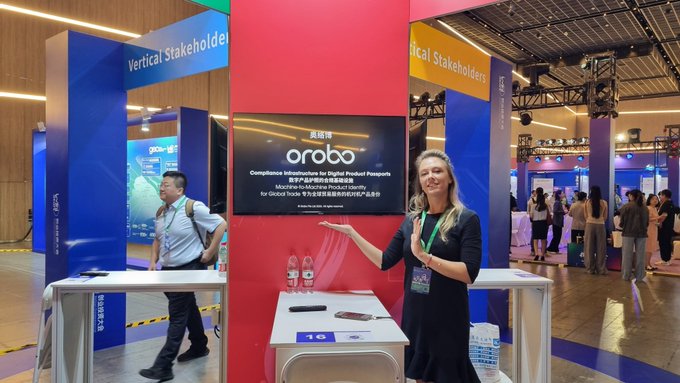 A woman stands at a trade show booth presenting for Orobo, a Chinese compliance infrastructure company for digital product passports, with the display noting "Modular Microservice Product Identity for Global Trade" and showing product samples on a table.