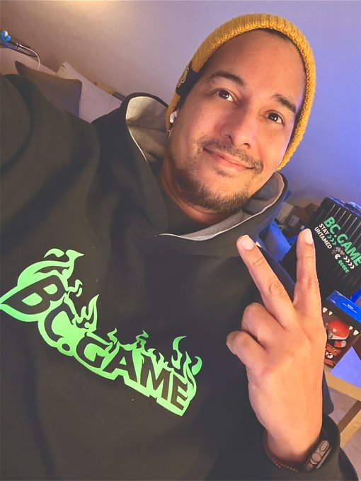 A man wearing a yellow beanie and a black hoodie with green "BC.GAME" flame-style lettering takes a selfie while flashing a peace sign, with a "BC.GAME" branded box visible on a shelf behind him—tying into the "GN fam" (good night family) post as a casual sign-off to his community or fans.