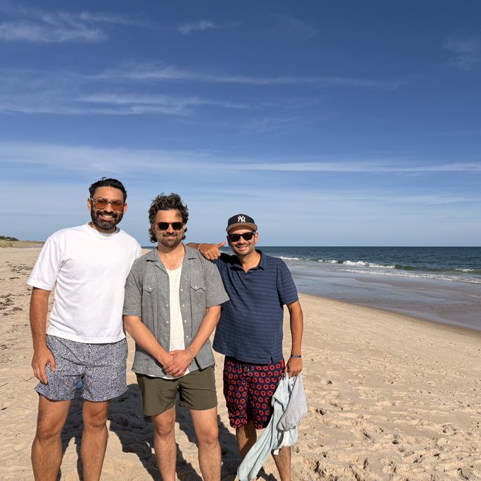 Three men pose together on a sandy beach under a bright blue sky, with the ocean visible in the background.