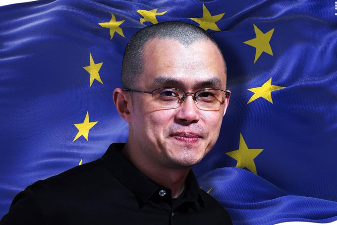 A man with short hair and glasses, wearing a black shirt, stands in front of a European Union flag featuring yellow stars on a blue background. The image accompanies a breaking news report where CZ comments on the EU's decision not to grant Binance a MiCA license, which he claims would restrict users from accessing top global liquidity.