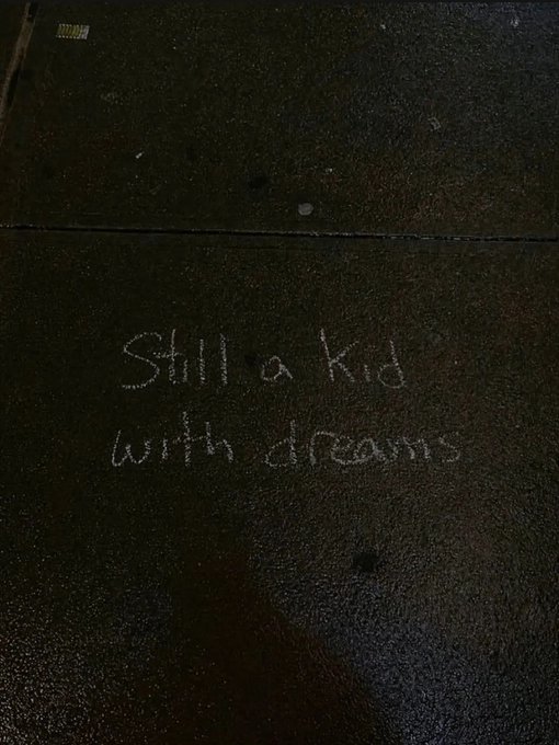 Chalk writing on a wet pavement that reads 'Still a Kid with dreams'.