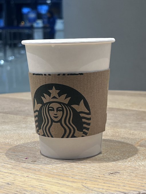 A white paper coffee cup with a brown cardboard sleeve displaying the Starbucks logo sits on a wooden table in what appears to be a café setting.