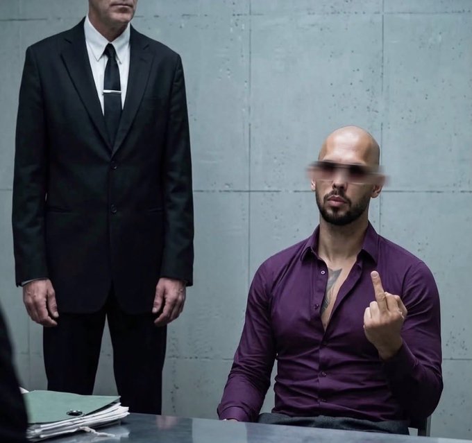 A cinematic scene of a seated bald man with a beard making an obscene gesture, while a tall figure in a black suit and tie stands behind him against a concrete wall.