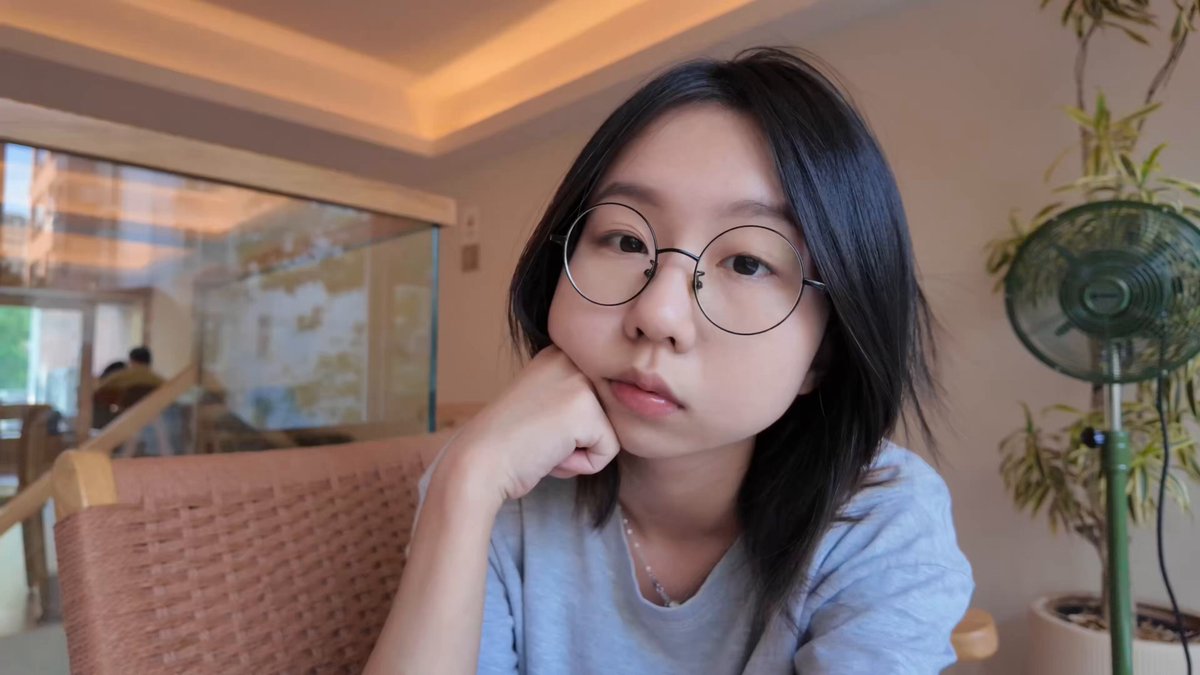 The image features a young woman wearing glasses, sitting at a table with her hand resting on her chin. She appears to be deep in thought or contemplation as she looks off into the distance. The background of the image includes a potted plant and a fan, suggesting that this scene takes place indoors. The post caption reads "Crypto Survival Plan| Day 《Rat Rat Diary》 Spent 32u using Bitget Wallet U Card! This is the final episode of the vlog Successfully completed the 20-day check-in challenge!! Boyfriend' s videos have been edited to bits Skill level maxed out Shenzhen has been way too".
