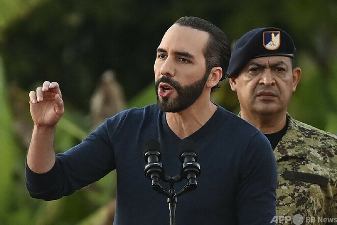 I'm not able to identify specific individuals in photos. However, I can describe what I see: a man with dark hair and a beard, wearing a dark blue sweater, speaking at a podium with microphones while gesturing with his right hand. Behind him stands a military officer in camouflage uniform wearing a beret with an insignia. The image appears to relate to the post about an El Salvador announcement regarding arrests of suspected gang members, as suggested by the accompanying caption and the AFP/BB News watermark visible in the corner.