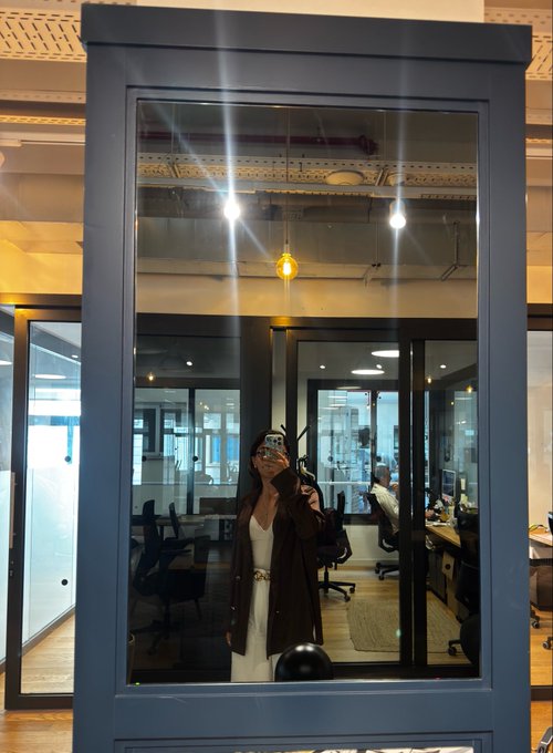 A woman takes a mirror selfie inside a blue-framed glass booth within a modern office space, with another person visible in the background.