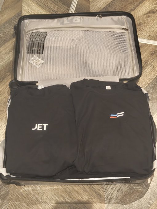 An open suitcase containing two neatly folded black t-shirts, one featuring the text 'JET' and the other displaying a small French flag logo.